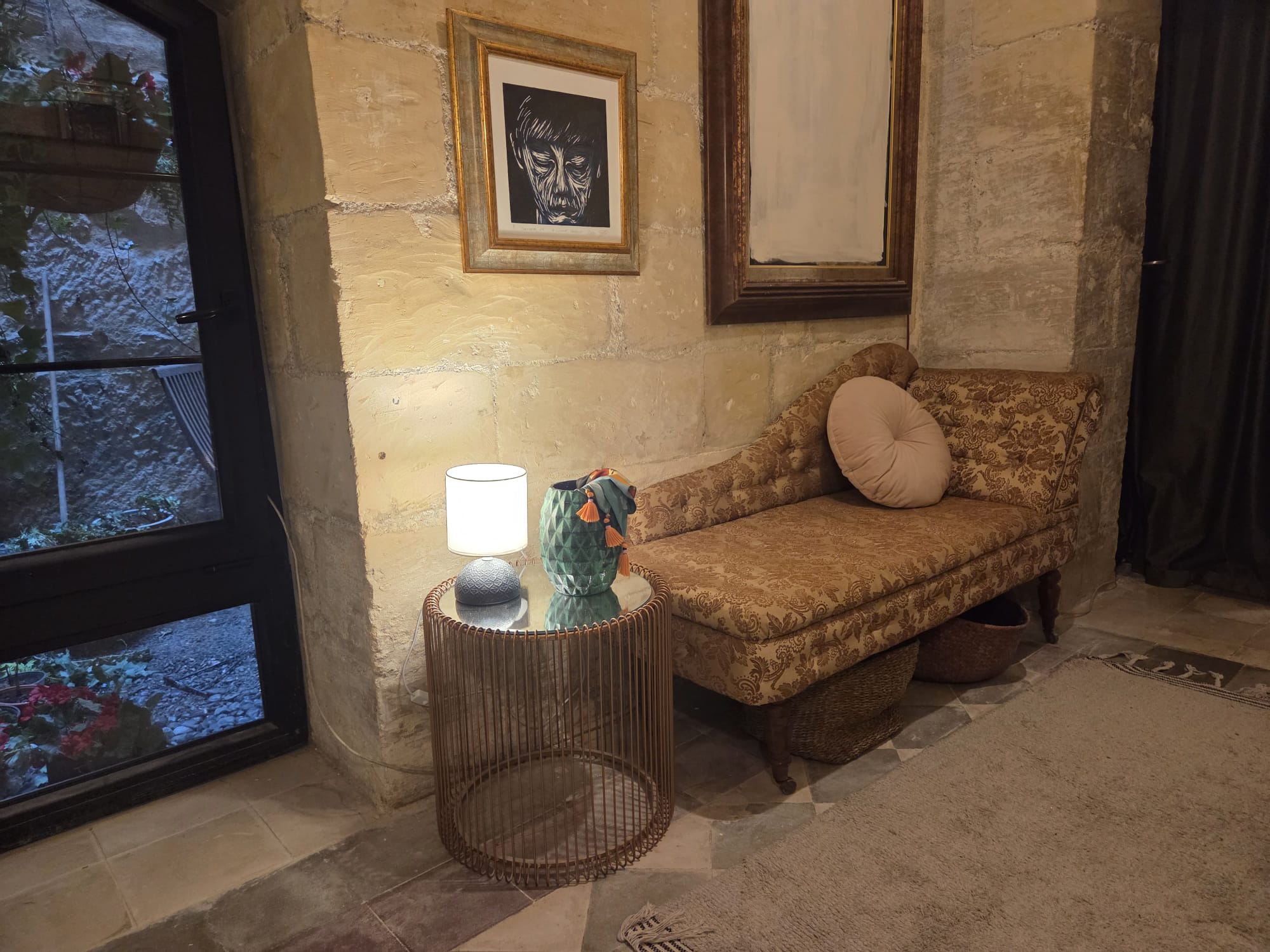 A chaise longue in gold brocade against a limestone wall, beside a black-framed glazed door to the courtyard. A wire side table holds a small lamp and a green faceted vase; above hang a framed linocut portrait and a tall mirror.
