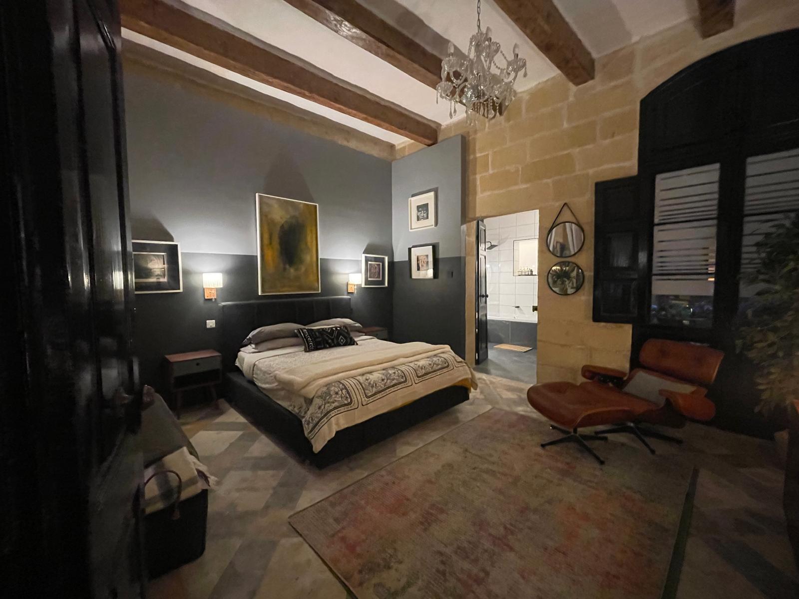 A bedroom with dark wood ceiling beams, half-limestone half-charcoal walls, a Venetian glass chandelier, gallery wall of small framed works, and a window bench set into a thick wall embrasure.
