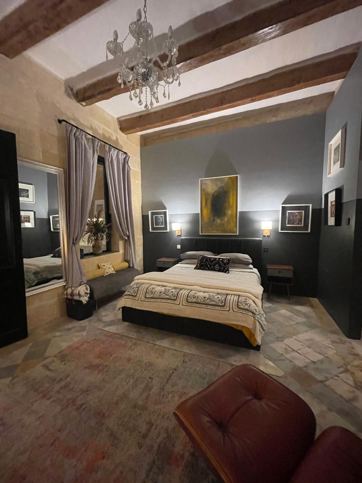 The bedroom seen from the opposite corner: chandelier, dark beamed ceiling, leather reading chair and red rug beside the bed.