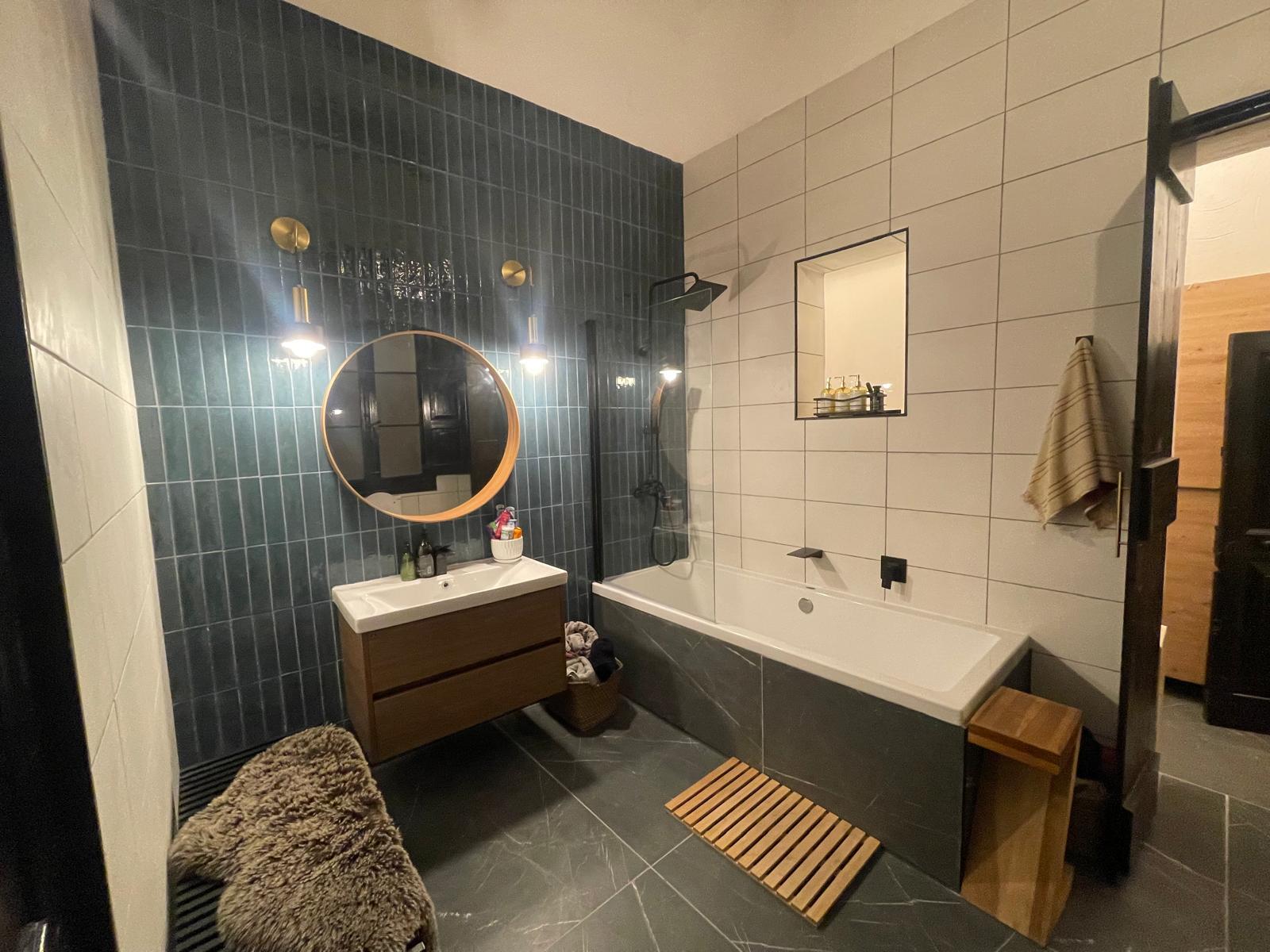A bathroom with dark blue-green tile walls, a freestanding tub, a round mirror, and white brick-pattern tile.