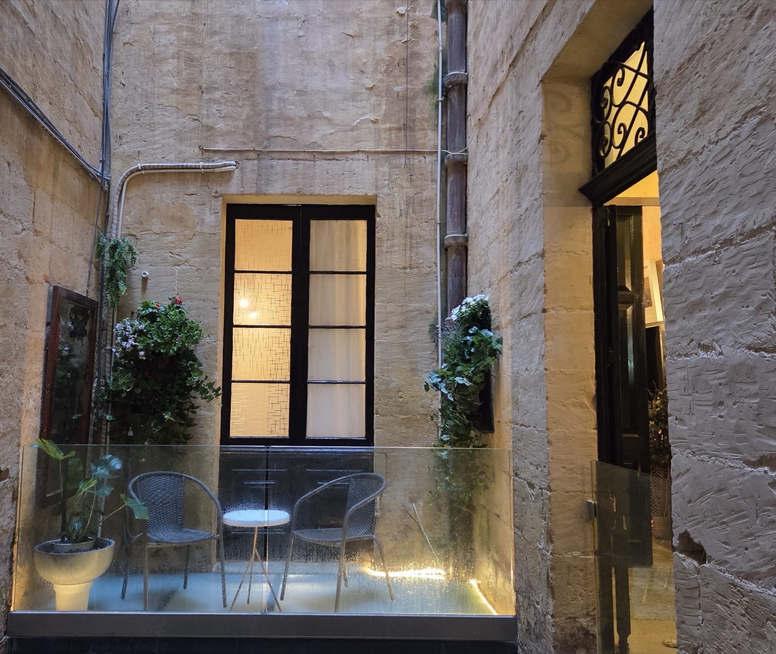 A small glass-floored courtyard terrace at dusk, set into a limestone light-well: a round bistro table and two woven chairs, potted plants and hanging flower baskets, a tall black-framed window glowing warm from within, and an open door beneath an ornate wrought-iron fanlight.